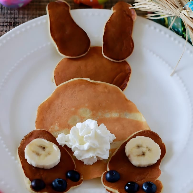 Bunny-shaped pancakes with whipped cream, banana slices, and blueberries on a white plate.