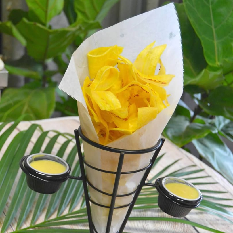 Crispy plantain chips in a paper cone with two dipping sauces, set against green foliage.