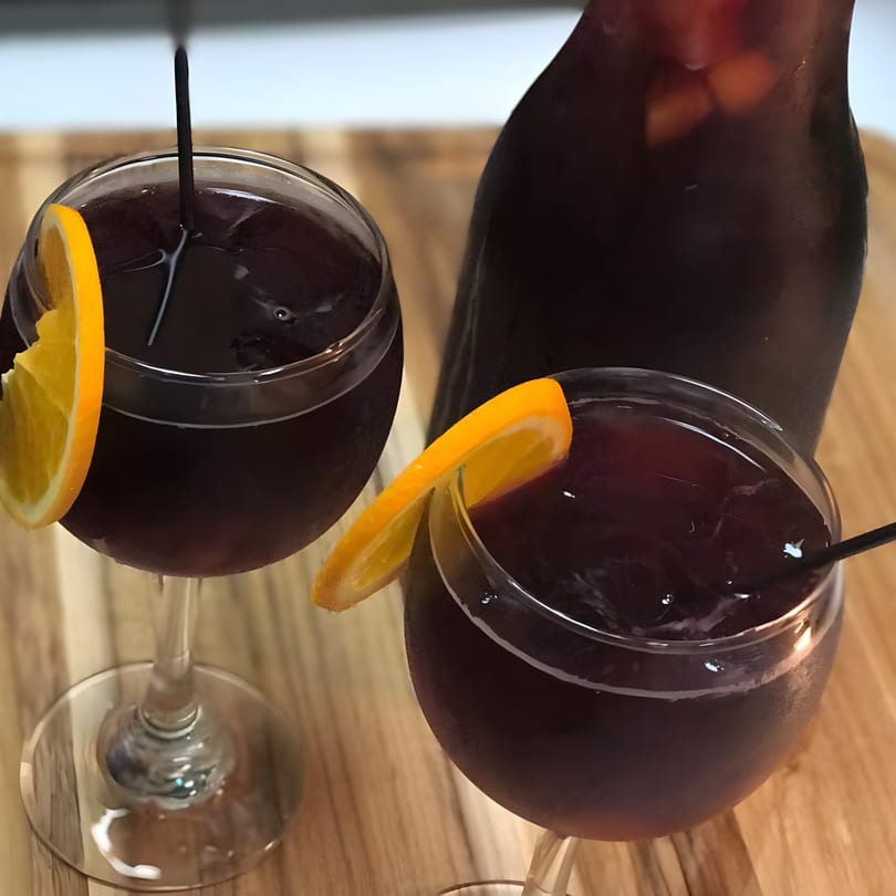 Two glasses of red sangria with orange slices and a pitcher on a wooden surface.