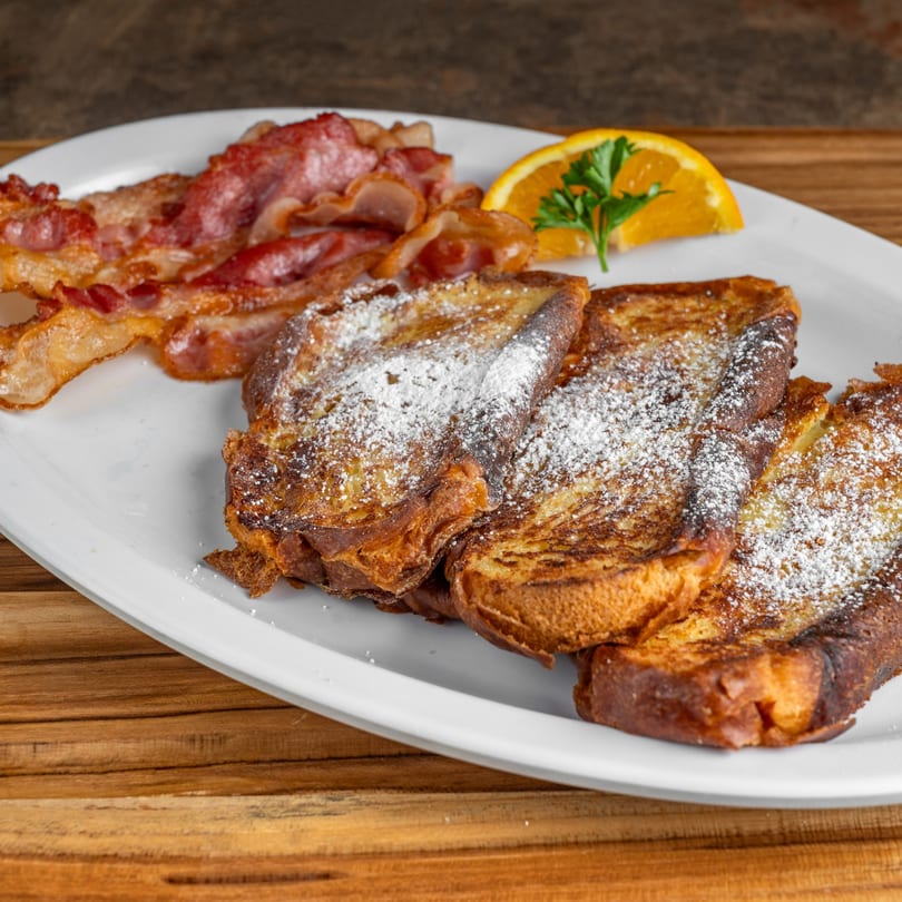 French toast with powdered sugar, crispy bacon, and an orange slice with parsley on a white plate.