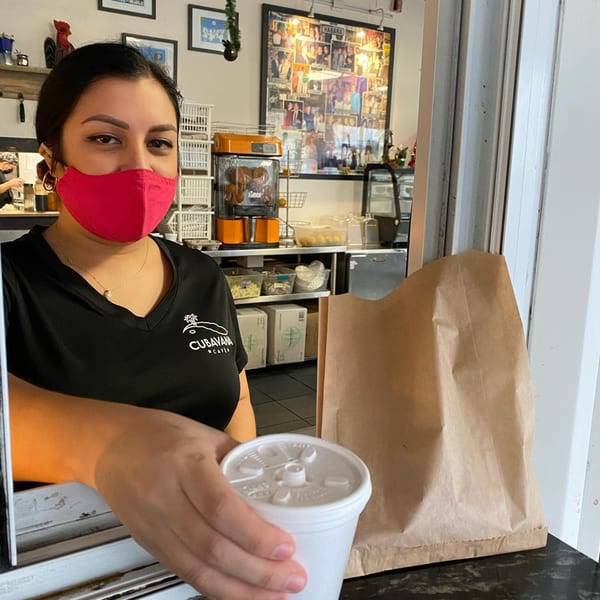A Cubavana Cafe employee in a red mask hands a drink and a brown paper bag through a window.