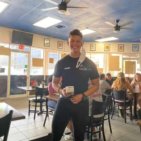 A smiling Cubavana Cafe employee in a black uniform holds a payment device in a bustling restaurant.
