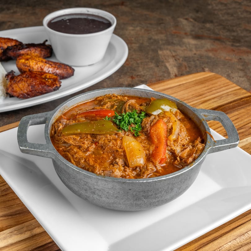 Cuban ropa vieja in a metal pot with bell peppers, served with black beans and plantains.